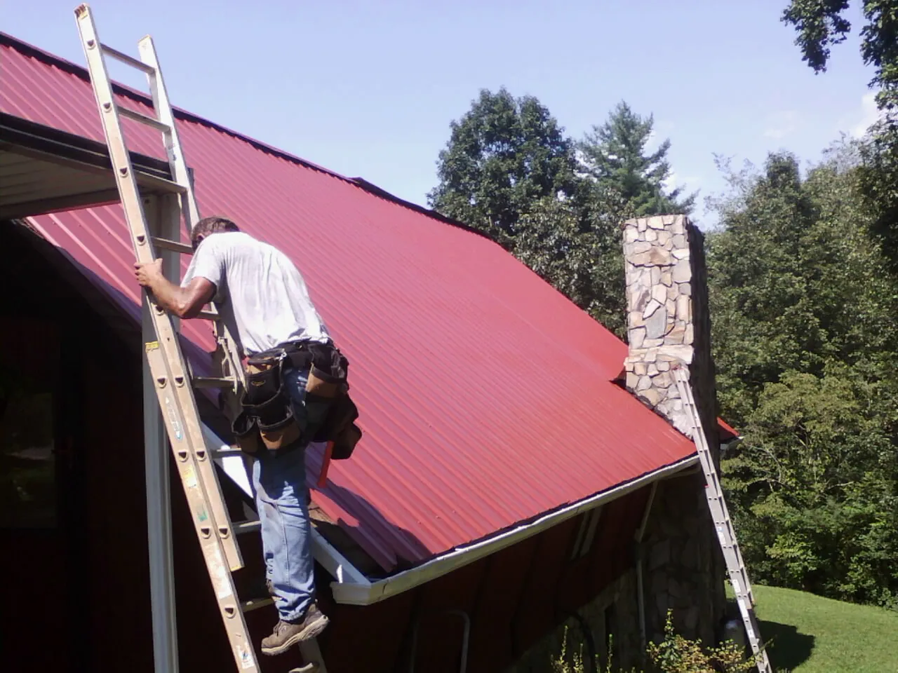 Roofer on a ladder beside a red metal roof and stone chimney — Soffit & Fascia Repair by Roofing Brothers in Shepherdsville